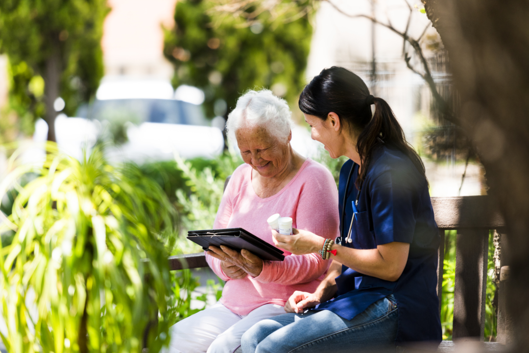 Caregiver and senior woman sitting outdoors. We make sure health needs are met with a high caregiver ratio.